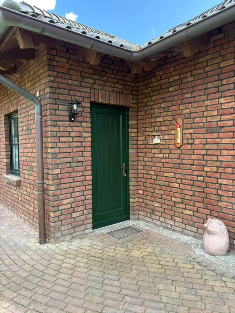 Dunkelgrüne Tür in einer Backsteinecke, links eine schwarze Laterne, rechts ein Willkommensschild und ein Holzthermometer, davor eine steinerne Igelfigur auf dem Pflaster