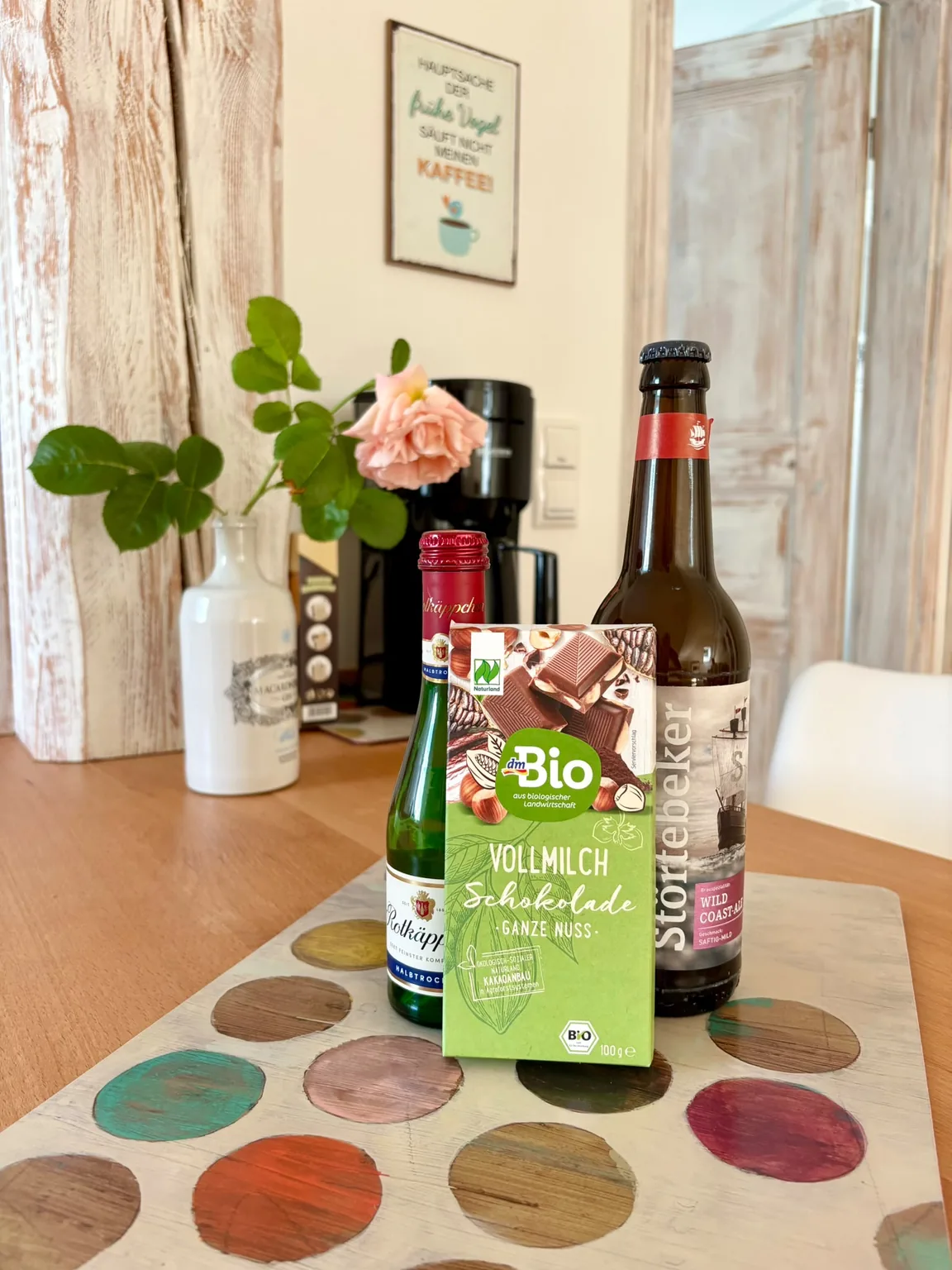 Sekt, Schokolade und Bier auf einem bunt bemalten Holzbrett, dahinter eine Rose in einer weißen Vase