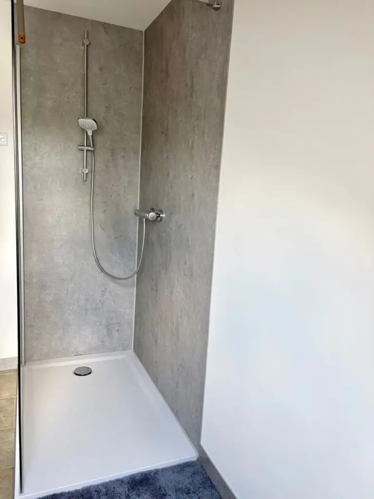 Duschwanne mit flachem Rand und Glaswand, Handbrause an einer Stange, Wandverkleidung in Betonoptik