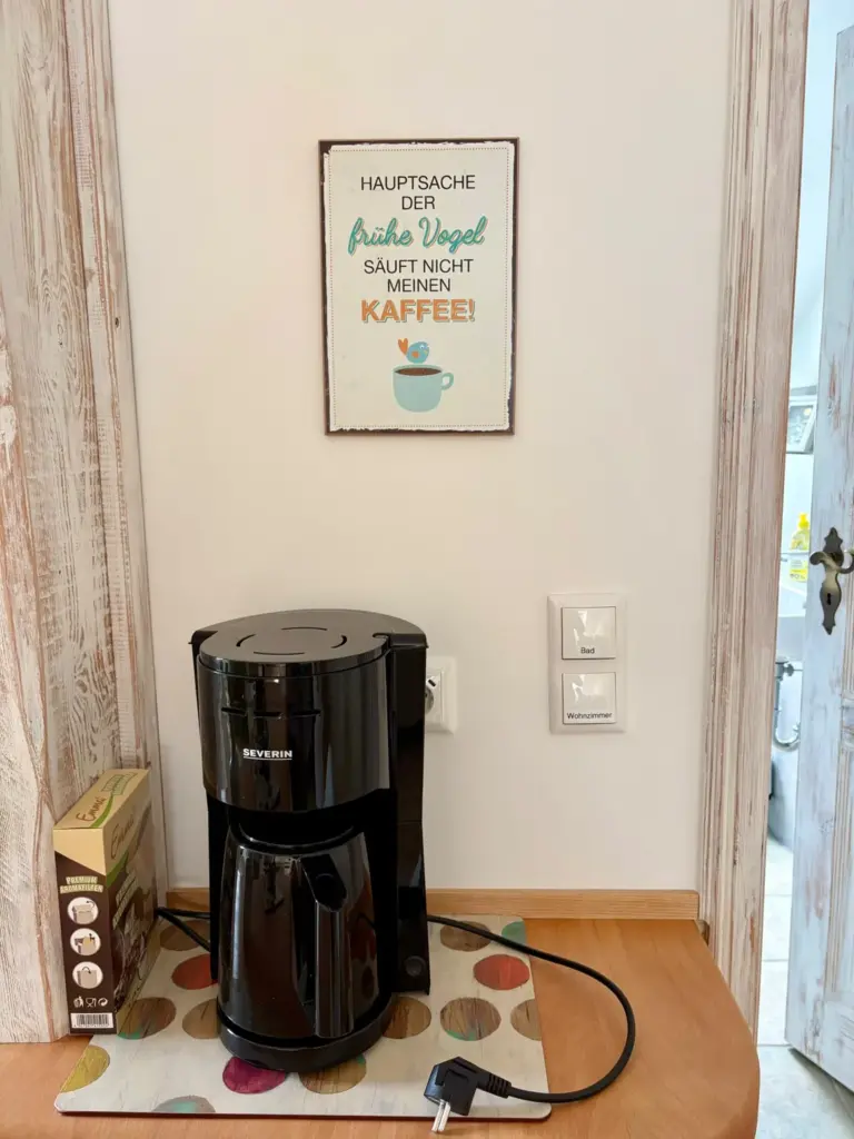 Filterkaffeemaschine mit Thermokanne auf einer Ablage im Flur, darüber ein Blechschild