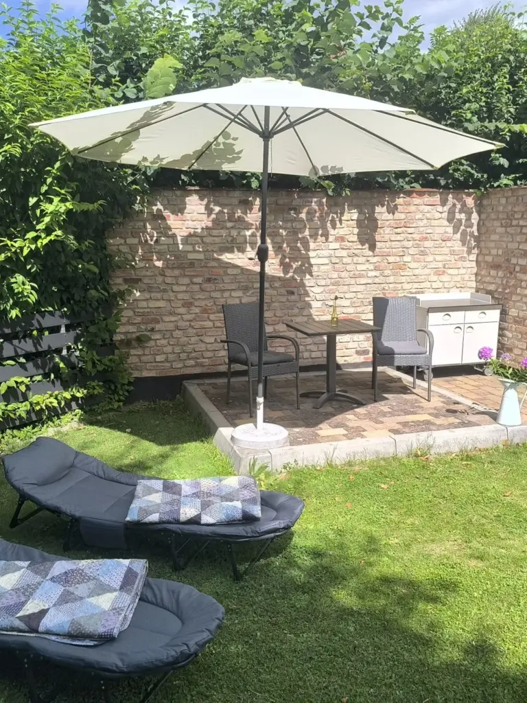 Sonnenschirm über Tisch und Sesseln vor einer alten Backsteinmauer, davor zwei Sonnenliegen auf dem Rasen
